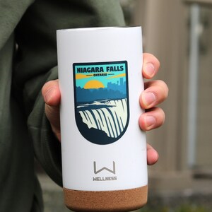 May include: White travel mug with a cork bottom. The mug has a sticker on it that says "Niagara Falls - Ontario" with a graphic of the falls and a sunset.