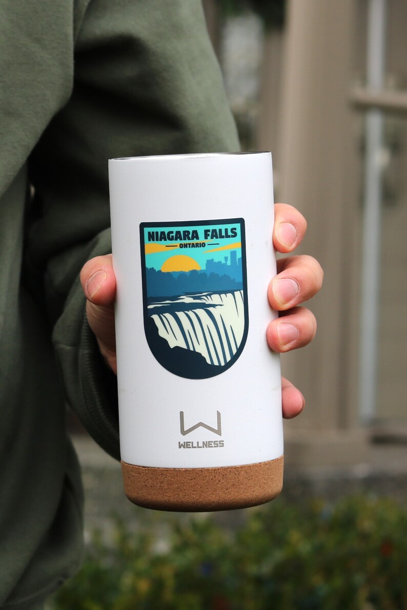 May include: White travel mug with a cork bottom. The mug has a sticker on it that says "Niagara Falls - Ontario" with a graphic of the falls and a sunset.