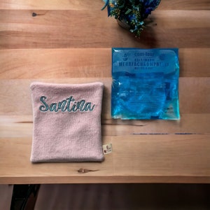 Könnte beinhalten: Ein rosa, personalisierter, bestickter Handwärmer mit dem Namen "Santina" in türkisfarbener Stickerei. Ein blauer Kalt-/Warmkompress mit dem Text "com-four KALT/WARM MEHRFACHKOMPRESSF HOT/COLD PAC" ist ebenfalls abgebildet.