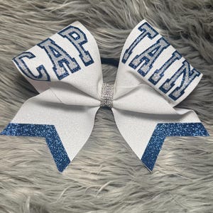 Puede incluir: Lazo de animadora blanco con la palabra "CAPTAIN" en letras plateadas con purpurina delineadas con purpurina azul. El lazo tiene detalles de purpurina azul y un centro de diamantes de imitación.