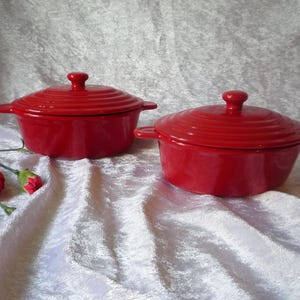 Puede incluir: Dos cazuelas de cerámica roja pequeñas con tapas. Cada plato tiene forma redonda con un asa a cada lado y un pomo en la tapa. Los platos están sobre una superficie blanca texturizada.