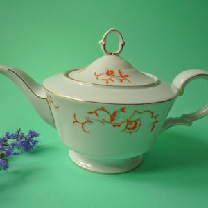 Op de afbeelding: Witte porseleinen theepot met oranje bloemmotieven en gouden randen. De theepot heeft een gebogen tuit en een afgeronde handgreep. De achtergrond is effen groen. Een takje paarse bloemen staat in de linker benedenhoek.