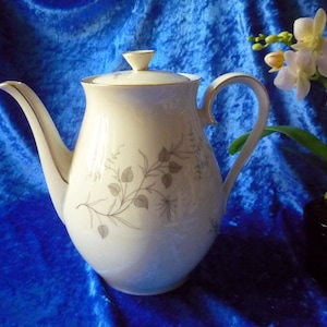Op de afbeelding: Een crèmekleurige porseleinen theepot met een delicaat grijs botanisch patroon van bladeren en ranken. De theepot heeft een bijpassend deksel met een kleine knop, een gebogen handvat en een slanke tuit, allemaal afgewerkt met een dunne gouden rand.