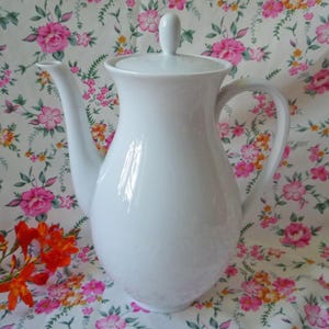 Può includere: Una caffettiera in ceramica bianca con coperchio e manico curvo. La caffettiera ha un lungo beccuccio e un corpo arrotondato. Lo sfondo è un motivo floreale con fiori rosa e arancioni.