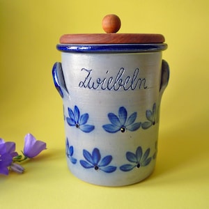 Peut inclure: Un pot de stockage cylindrique en céramique avec une finition gris clair, doté de deux anses latérales et d'un couvercle en bois avec un bouton rond. Le pot est décoré de motifs floraux bleus peints à la main et du mot Zwiebeln en bleu.