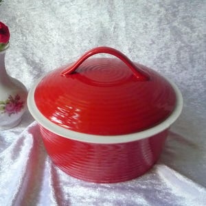 Può includere: Una pirofila in ceramica rossa con bordo bianco e coperchio abbinato. Il coperchio ha una maniglia e un design strutturato a cerchi concentrici. A sinistra, un piccolo vaso bianco con dettagli floreali e un fiore rosso.