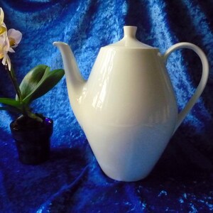 Könnte beinhalten: Eine weiße Porzellan-Teekanne mit gebogenem Griff und Deckel, vor blauem Hintergrund. Links von der Teekanne befindet sich eine kleine Topf-Orchidee mit gelben und weißen Blüten. Die Teekanne hat ein Fassungsvermögen von ca. 1,5 Liter.