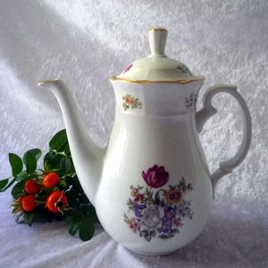 Può includere: Caffettiera in ceramica bianca con coperchio, decorata con motivi floreali viola, rosa e gialli. La caffettiera ha un manico curvo e un bordo dorato. Un rametto di bacche rosse e foglie verdi è accanto.
