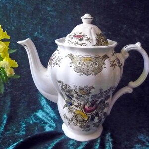 Op de afbeelding: Witte keramische theepot met deksel, versierd met bloemen- en krulpatronen in groen, geel en roze. De theepot heeft een gebogen tuit en handvat. Een klein boeket gele bloemen staat op de achtergrond.