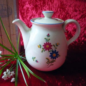 Op de afbeelding: Crèmekleurige keramische theepot met een deksel, versierd met kleurrijke bloempatronen. De theepot heeft een gebogen tuit en handvat, en een blauwe rand. De achtergrond is een rode fluwelen stof.