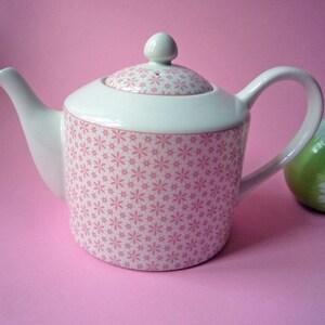 Op de afbeelding: Een witte keramische theepot met een herhalend patroon van kleine roze bloemetjes op de romp en het deksel. De theepot heeft een gebogen handvat, een taps toelopende tuit en een rond deksel met een kleine knop bovenop, tegen een roze achtergrond.