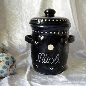 Puede incluir: Un tarro de cerámica azul oscuro con tapa, asas y la palabra "Müsli" en escritura blanca. El tarro está decorado con puntos blancos. Dos esferas decorativas con diseños florales azules y blancos en el fondo.
