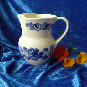 Op de afbeelding: Witte keramische kan met blauwe bloemmotieven. De kan heeft een gebogen handvat en een schenktuit. Oranje en gele bloemen zijn naast de kan geplaatst op een blauwe fluwelen stof.