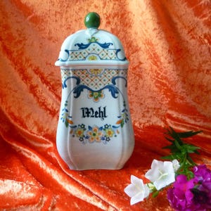 Puede incluir: Un recipiente de cerámica blanca con tapa extraíble y un pomo verde. El recipiente está decorado con patrones florales y de celosía en azul, amarillo y verde. La palabra Mehl está impresa en letra gótica negra en la parte frontal.