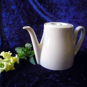 Może przedstawiać: Biały ceramiczny czajnik z pokrywką i dzióbkiem. Czajnik stoi na niebieskim aksamitnym podłożu.