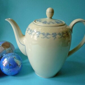 Può includere: Teiera in porcellana color crema con coperchio e manico curvo. La teiera presenta un delicato motivo floreale blu sul bordo e sul coperchio. Tre sfere decorative in vetro blu e iridescente sono in primo piano.