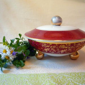 May include: A decorative ceramic serving dish with a lid. The dish is red with gold floral patterns and a white rim. It has a gold knob on top and rests on three gold feet. A small bouquet of flowers is next to the dish.