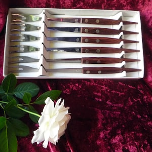 Puede incluir: Un juego de cubiertos vintage en una caja blanca, con tenedores y cuchillos de acero inoxidable con mangos de madera oscura. El juego se exhibe sobre una superficie de terciopelo rojo oscuro, con una rosa blanca y hojas verdes en primer plano.