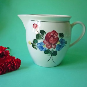 Könnte beinhalten: Ein weißer Keramikkrug mit einem handgemalten Blumenmotiv, bestehend aus einer großen roten Rose in der Mitte, kleineren blauen Blüten und grünen Blättern. Der Krug verfügt über einen geschwungenen Henkel, einen Ausguss und einen grünen Rand.
