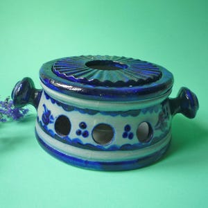 Może przedstawiać: Niebiesko-biały ceramiczny garnek z pokrywką. Okrągły garnek ma dwa uchwyty i kilka okrągłych otworów. Pokrywka ma teksturowaną, żebrowaną powierzchnię. Ozdobiony motywami kwiatowymi. Artykuł dekoracyjny.