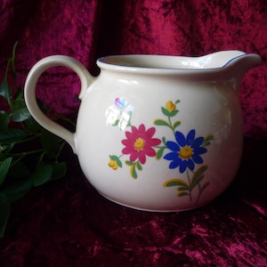 Può includere: Brocca in ceramica color crema con corpo arrotondato e manico. Decorata con motivi floreali rosa e blu e un bordo blu. Un pezzo classico per servire bevande.