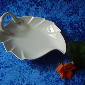 Può includere: Un piatto da portata in ceramica bianca a forma di foglia con manico. Il piatto ha una finitura liscia e lucida ed è posto su uno sfondo blu. Un fiore arancione e una foglia verde sono accanto al piatto.