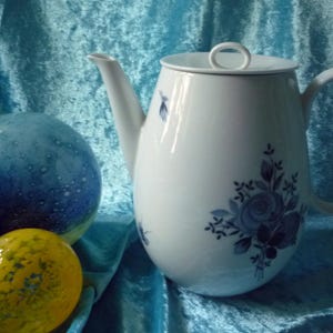 Puede incluir: Tetera de cerámica blanca con tapa y asa, adornada con diseños florales azules. La tetera se encuentra sobre un fondo azul con esferas decorativas de vidrio en azul y amarillo.