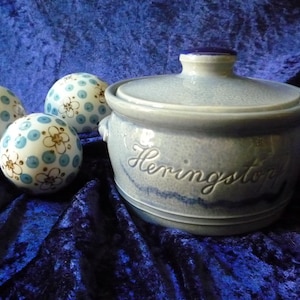 Op de afbeelding: Een lichtblauwe keramische pot met de inscriptie "Heringston" en een deksel, vergezeld van drie decoratieve bollen. De bollen zijn wit met blauwe stippen en bloemmotieven. De items staan op een donkerblauwe fluwelen ondergrond.