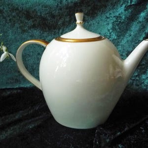 Può includere: Una teiera in porcellana bianca con corpo arrotondato, manico curvo e beccuccio. Il coperchio e il bordo sono rifiniti con finiture dorate. Un piccolo rametto di fiori bianchi è visibile a sinistra della teiera.
