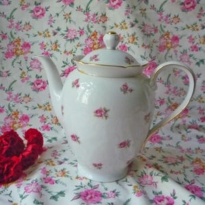 May include: White porcelain teapot with a lid, decorated with small pink rose patterns and gold trim. The teapot has a curved handle and a spout. The background features a floral pattern with pink and yellow roses.