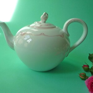 Puede incluir: Una tetera de porcelana blanca con cuerpo redondeado, asa curva y boquilla. La tapa está rematada con un pequeño adorno. Delicados diseños florales rosas adornan la tetera. Una rosa rosa y capullos están en primer plano.
