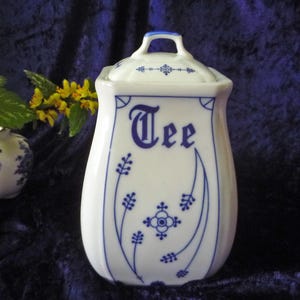 Könnte beinhalten: Weiße Keramik-Teedose mit blau-weißem Blumenmuster. Die Dose hat einen Deckel mit einem Knopf und das Wort "Tee" auf der Vorderseite.