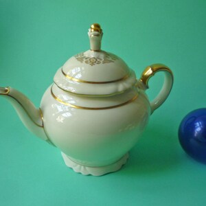 May include: A white porcelain teapot with gold trim and a matching lid. The teapot has a rounded shape and a curved spout. A blue glass ball sits next to the teapot. The teapot is likely for serving tea.