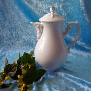 Könnte beinhalten: Eine weiße Porzellan-Kaffeekanne mit Deckel, Ausguss und verziertem Griff. Die Kanne steht vor einem blauen Hintergrund mit einem Zweig gelber Blumen und grünen Blättern.