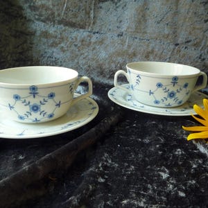 Può includere: Due tazze da zuppa in porcellana bianca con piattini abbinati. Ogni pezzo è decorato con un motivo floreale blu. Le tazze hanno due manici. Un fiore giallo e foglie verdi sono sullo sfondo.