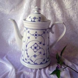 Op de afbeelding: Witte keramische koffiepot met een deksel, versierd met blauwe bloempatronen. De pot heeft een gebogen tuit en een handvat. Het ontwerp omvat blauwe bloemen en geometrische vormen. De pot staat op een wit oppervlak.
