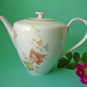Op de afbeelding: Een witte porseleinen theepot met een delicaat geschilderd patroon van herfstbladeren in tinten oranje, geel en groen. De theepot heeft een gebogen handvat, een taps toelopende tuit en een bijpassend deksel met een goudkleurige rand en knop.