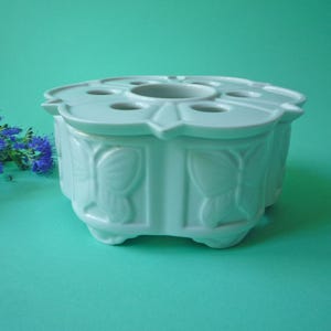 Może przedstawiać: Jasnoniebieski ceramiczny podgrzewacz do herbaty z kwiatowym wzorem. Podgrzewacz ma okrągłą górę z wieloma otworami i wzorem motyla po bokach. Podgrzewacz do herbaty stoi na małych nóżkach.