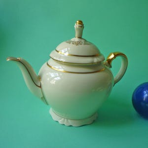 Può includere: Teiera in porcellana color crema con finiture dorate. La teiera presenta un corpo arrotondato, un beccuccio curvo e un manico dorato. Il coperchio ha un pomello dorato e dettagli decorativi. La teiera poggia su una base smerlata.