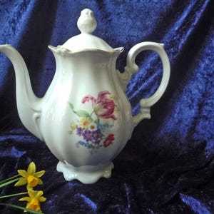 Op de afbeelding: Een witte porseleinen theepot met een geschulpte rand en een deksel met een bloemenprint van roze, gele en paarse bloemen. De theepot heeft een gebogen handvat, een lange tuit en een geribbeld ontwerp met een gouden rand op het deksel.
