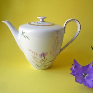 Puede incluir: Tetera de porcelana blanca con asa curva, boquilla y tapa. La tetera está decorada con diseños florales y de trigo en rosa, verde y dorado. Un racimo de campanillas moradas se encuentra a la derecha de la tetera sobre un fondo amarillo.