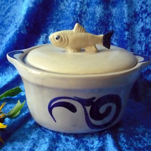 Op de afbeelding: Een keramische ovenschaal met een deksel, met een visvormige handgreep. De schaal is lichtblauw met donkerblauwe wervelontwerpen en randen. De vishandgreep is beige met een zwarte staart.