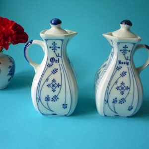 Peut inclure: Deux burettes en porcelaine blanche avec des motifs floraux bleus et des couvercles à pointe bleue, l'une étiquetée Oel et l'autre Essig, chacune dotée d'une anse et d'un bec. À côté se trouve un petit vase blanc à motifs bleus avec des fleurs rouges.