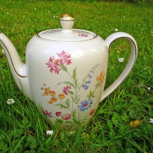 Puede incluir: Una tetera de porcelana blanca con un diseño floral con flores rosas, azules y amarillas. La tetera tiene un asa y ribetes dorados, y un pomo dorado en la tapa. La tetera está sobre hierba verde.