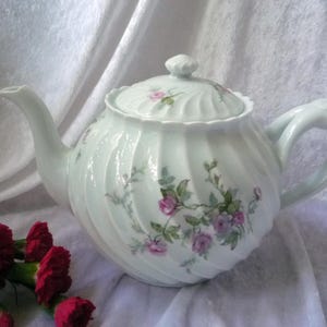 Op de afbeelding: Een witte porseleinen theepot met een deksel, met een spiraalvormig ontwerp en bloemaccenten in roze en groen. De theepot heeft een gebogen handvat en tuit. Rode bloemen staan op de voorgrond.
