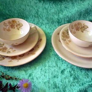 Peut inclure: Deux services à thé vintage avec des motifs floraux. Chaque ensemble comprend une tasse à thé, une soucoupe et une petite assiette. Les tasses et les assiettes ont un fond crème avec des bordures dorées et des motifs floraux dans les tons de marron et de jaune.