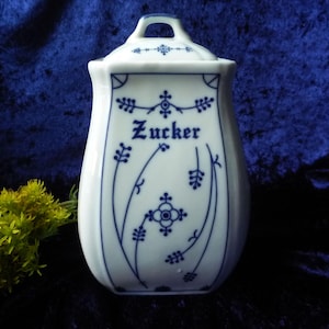 Puede incluir: Azucarero de cerámica blanca con tapa y asa, adornado con motivos florales azules y la palabra "Zucker". El recipiente tiene forma curvada y está sobre un fondo azul oscuro.
