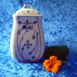 Puede incluir: Un recipiente de cerámica blanca para harina con tapa, adornado con patrones florales azules y la palabra alemana "Mehl". Una flor naranja y una hoja verde descansan junto al recipiente sobre un fondo azul.