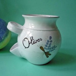 Może przedstawiać: Biały ceramiczny słoik na oliwki z bocznym wylewem i kwiatowym wzorem. Słoik jest oznaczony napisem "Olives" czarnymi literami.
