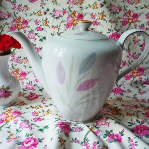 Puede incluir: Una tetera de porcelana blanca con un delicado patrón de hojas en tonos pastel rosa, morado y azul, rematada con un remate dorado. A su lado hay un pequeño jarrón de porcelana blanca decorado con un motivo floral de rosas rosas.